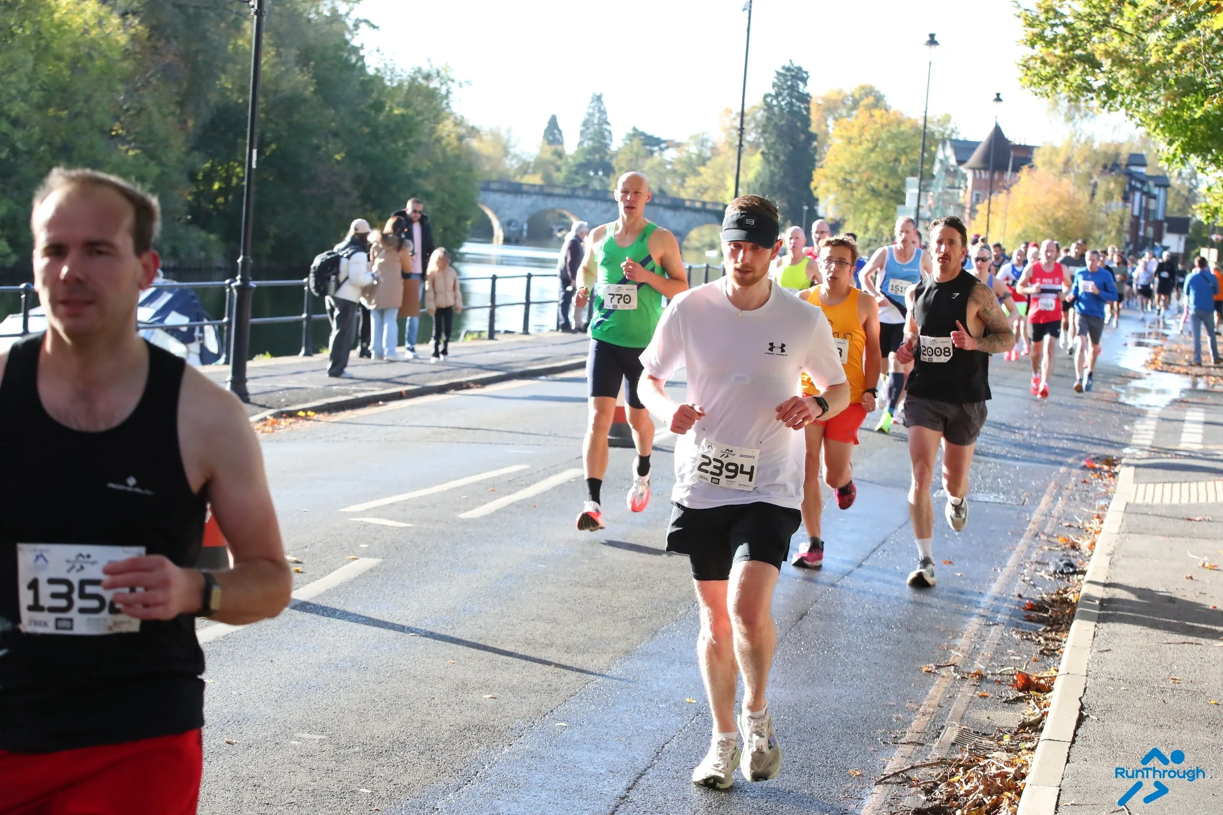 A group of marathon runners participating in a race on a city street next to a river, with trees and houses in the background on a sunny day.