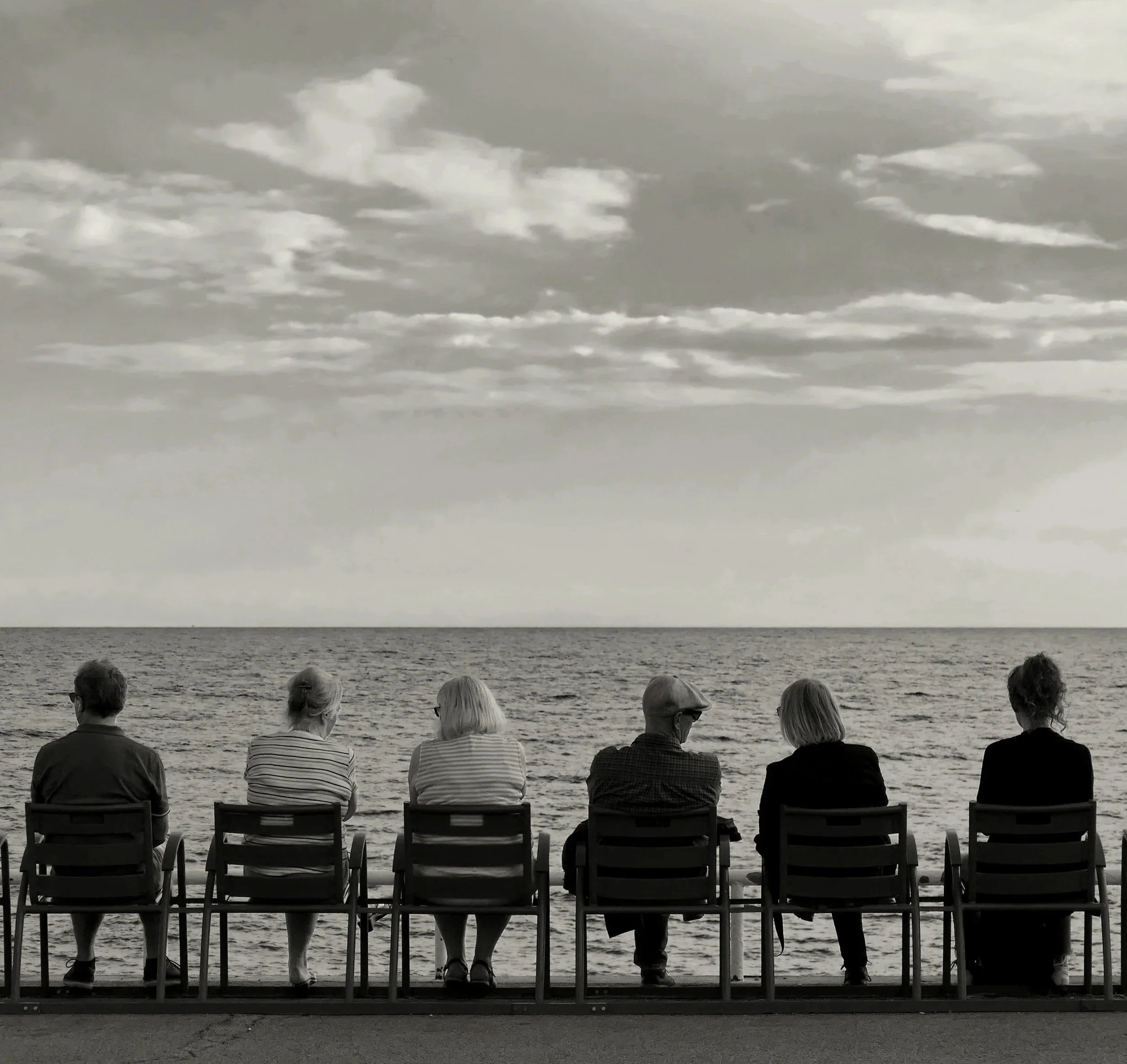 Six personnes sont assises sur des bancs face à la mer, en regardant l'horizon, dans une scène en noir et blanc.