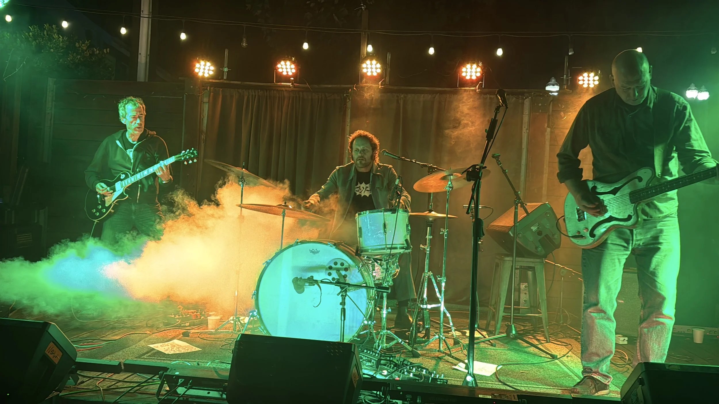 Suite G Studio's Grooveworks Team building three musicians perform on stage with colorful lighting and fog effects. One plays an electric guitar on the left, the second plays drums in the center, and the third plays an electric guitar on the right.