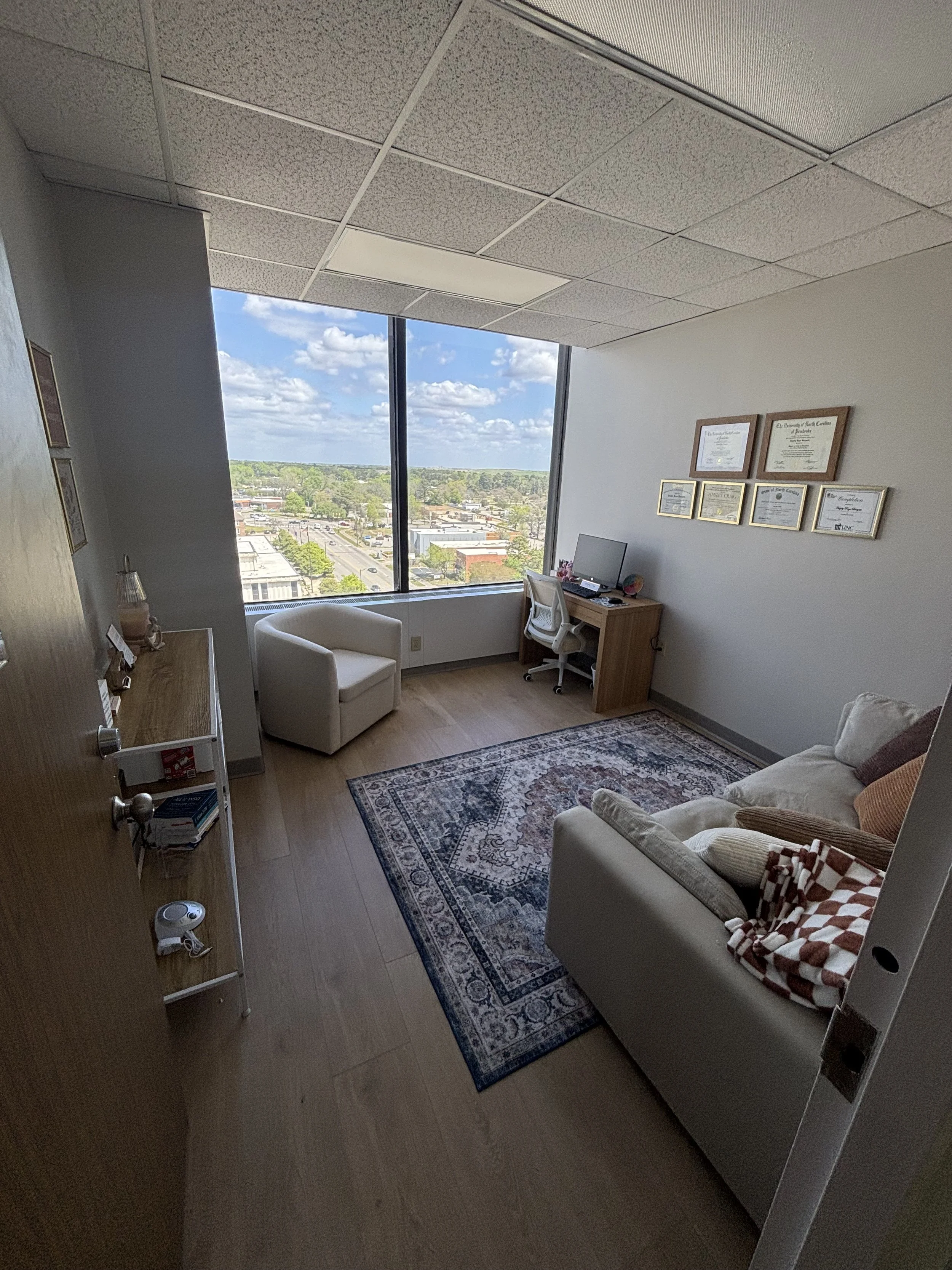 A Therapy room with a large window showing a view of a cityscape and blue sky, furnished with a white armchair, a wooden desk with a computer, framed certificates on the wall, a patterned rug, and a beige sofa with pillows.