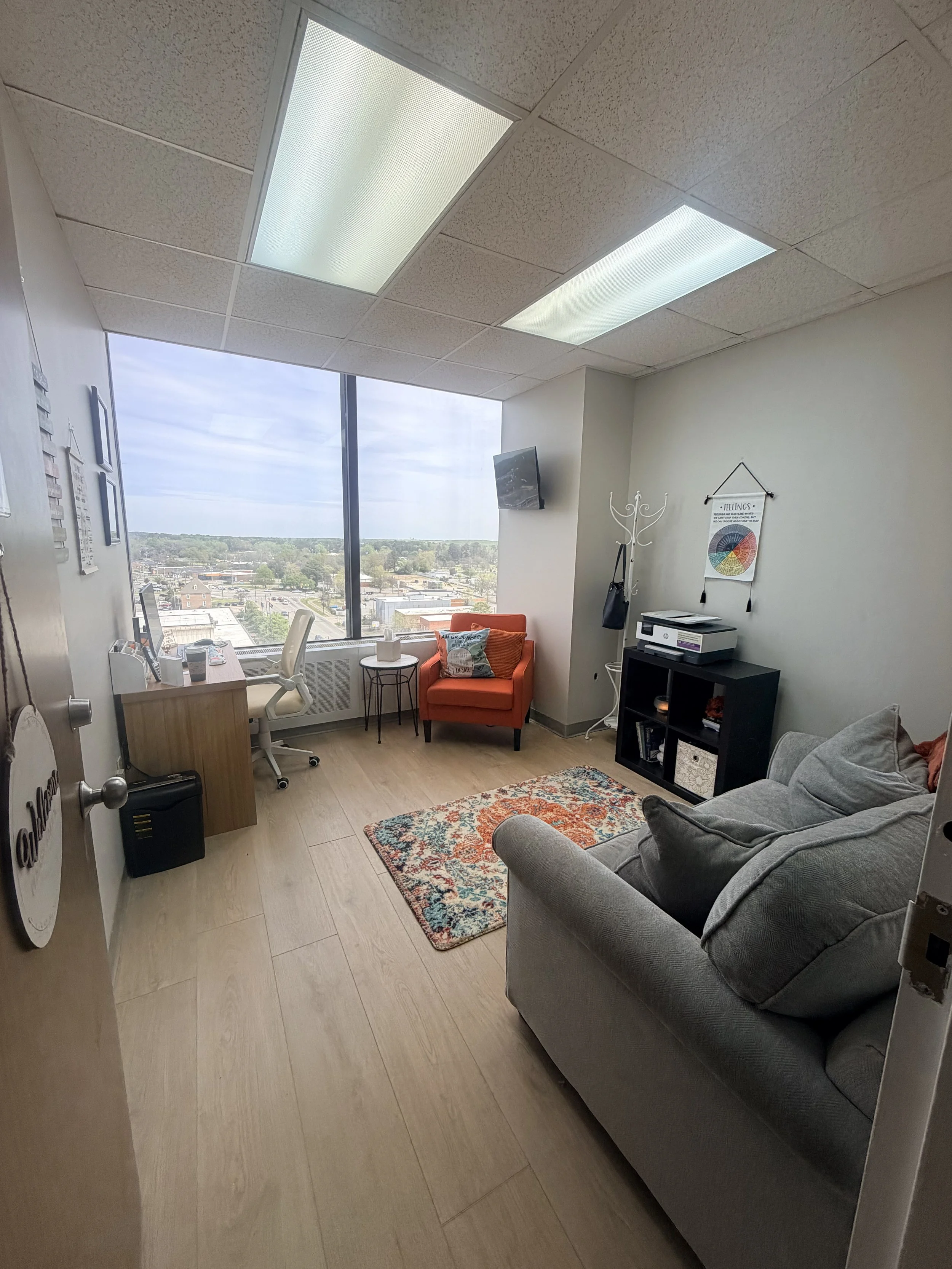 A therapy room with a large window, orange armchair, small side table, gray sofa, patterned rug, black bookshelf with binders, wall decorations, coat rack, TV on wall, ceiling lights.