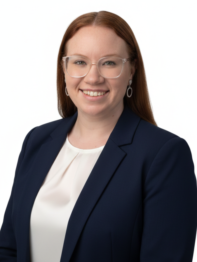 A woman with long red hair, glasses, and earrings wearing a navy blazer and white blouse posing against a white background.