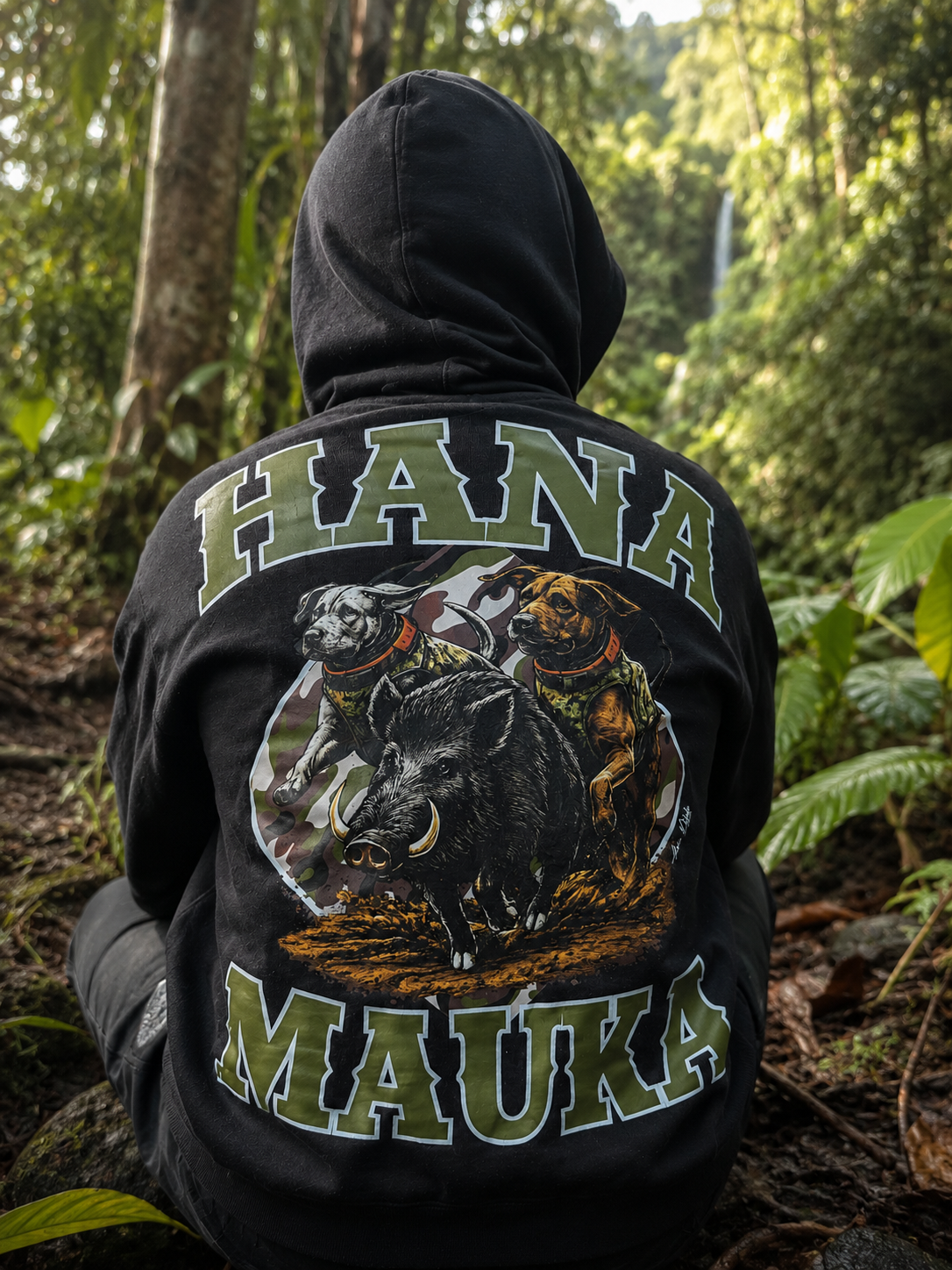 Person wearing a black hoodie with a graphic of two dogs and a boar, with text in Hawaiian on the back, sitting in a wooded area