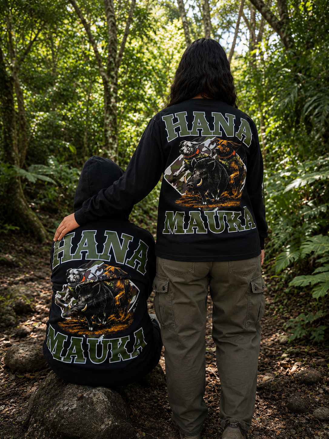 Two individuals wearing black jackets with a graphic of a boar and two dogs on the back, the words 'HANA MAUKA' printed on the jackets, standing and crouching in a dense, green forest.