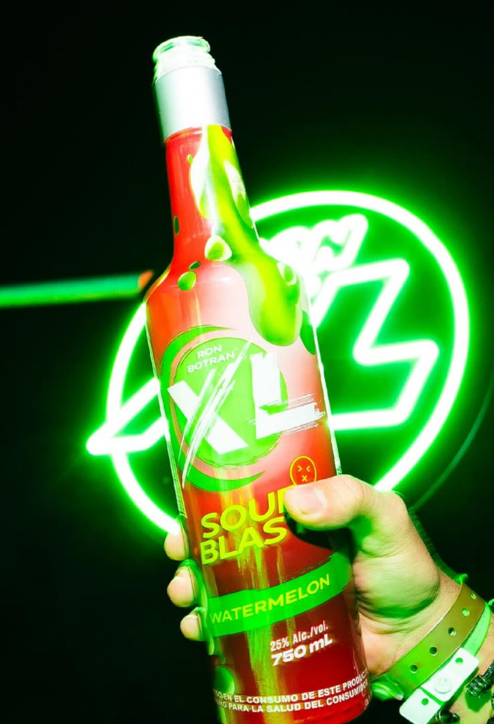 Hand holding a 750ml bottle of X-L Sour Blast Watermelon alcoholic beverage in front of a green neon sign.