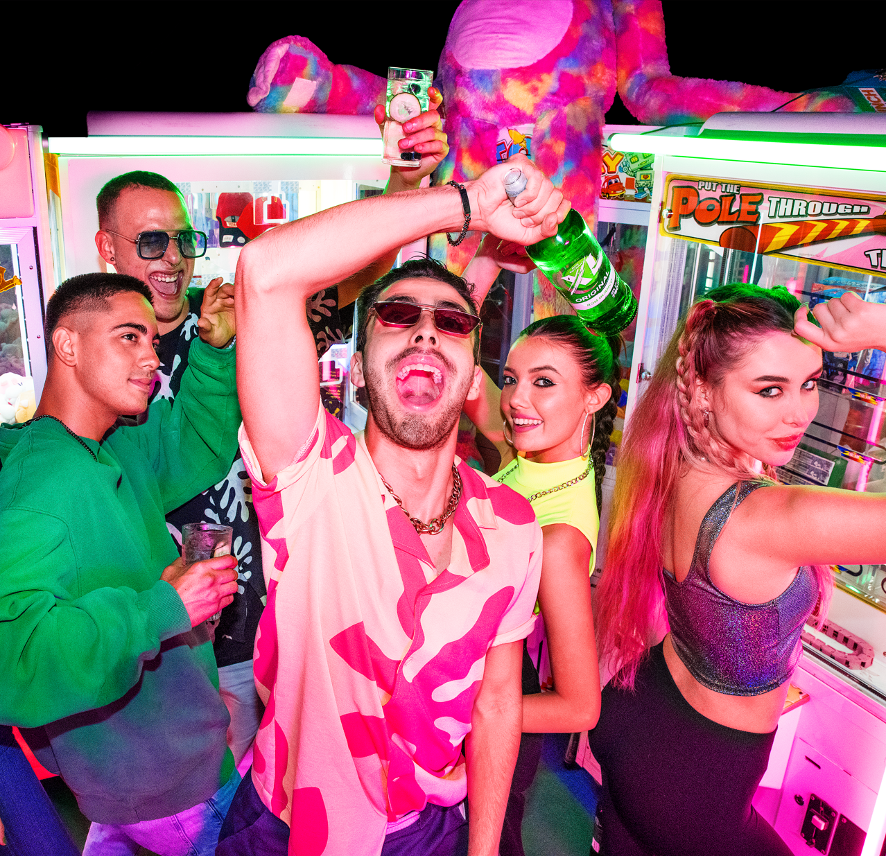Group of young people celebrating at an arcade, some wearing sunglasses, holding drinks, and dancing in front of claw machine games with neon lights.