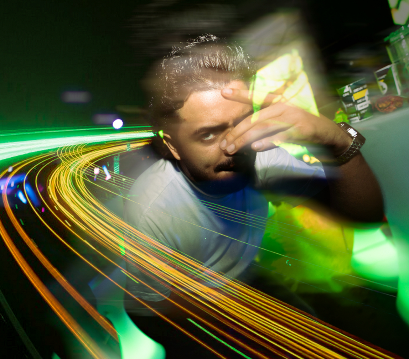A young man with dark hair and a watch on his left wrist is sitting at a table, resting his face on his right hand, looking into the camera. The photo features colorful light trails and streaks of neon green, yellow, and blue, creating a dynamic, futuristic effect, with shelves of snack food items visible in the background.