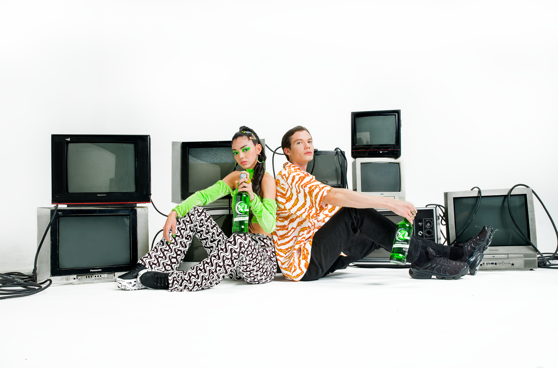 Two young people sitting on the floor surrounded by vintage television sets, holding bottles of green soda, with a plain white background.