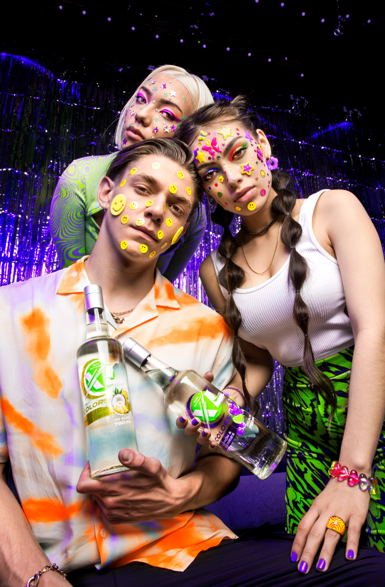 Three young people at a party with colorful face and body decorations, holding bottles of clear spirits, and standing in front of a purple tinsel backdrop.