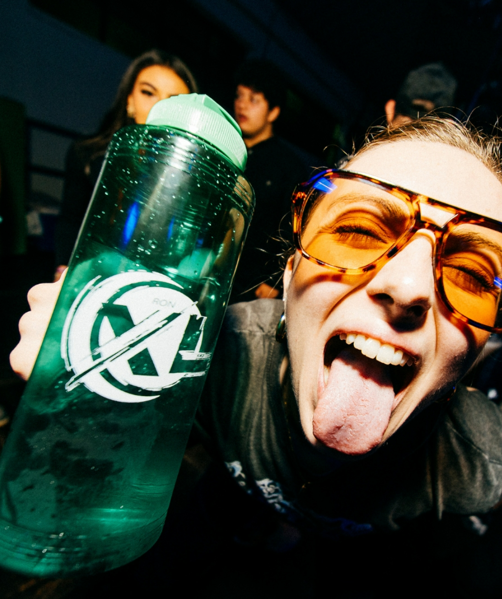 A woman at a party making a silly face with her tongue out, wearing orange-tinted sunglasses, holding a green water bottle with a logo, with two people blurred in the background.