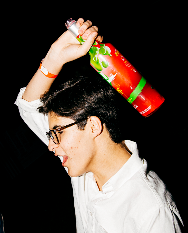 A young man with glasses and dark hair wearing a white shirt, holding a bottle of strawberry-flavored sparkling water above his head, appearing to pour or play with it against a black background.