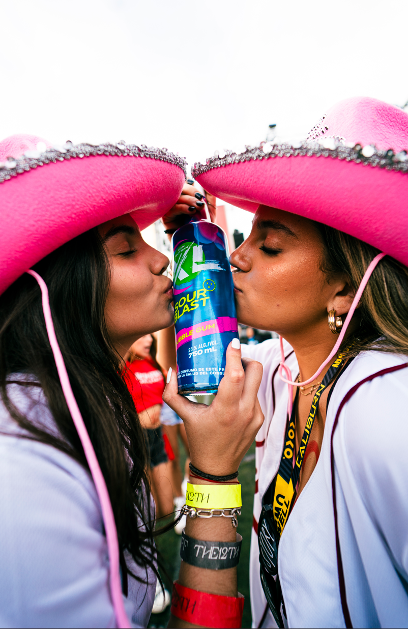 Two women wearing pink sombreros and white shirts sharing a drink from a can at an outdoor event.