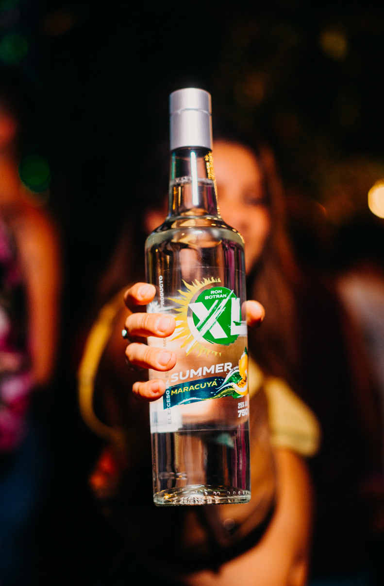 Close-up of a woman holding a bottle of alcohol with a colorful label that says 'Ron Botran X' and 'Summer Maracuyá' in a lively, nighttime setting.