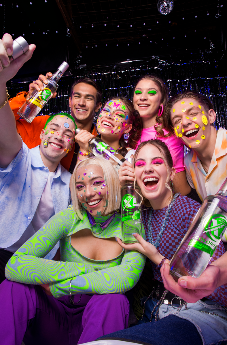 Group of young people celebrating at a party, smiling and holding bottles of alcohol, with colorful decorations and neon makeup.