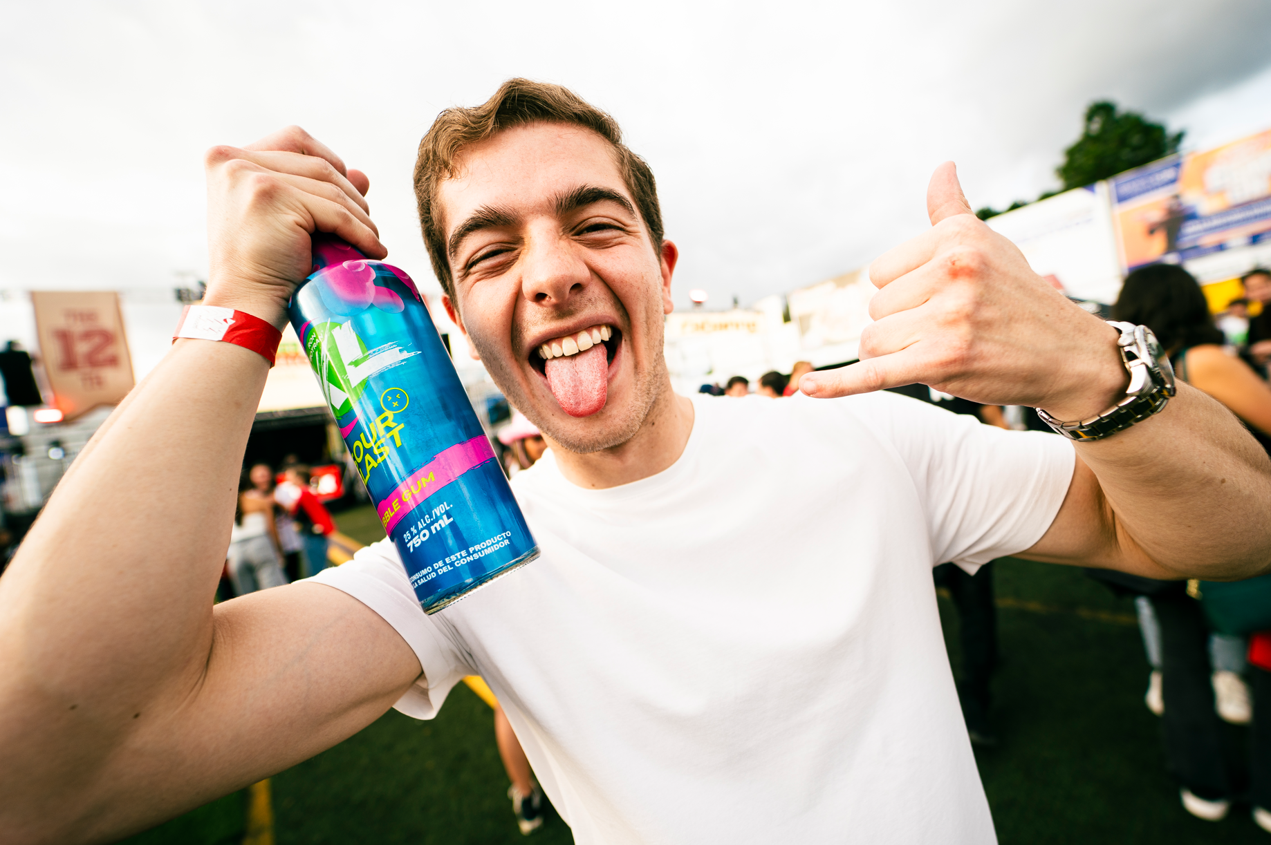 A young man at an outdoor event holding a colorful can of sour blast gum, sticking his tongue out, and making a shaka hand gesture, with a lively crowd in the background.