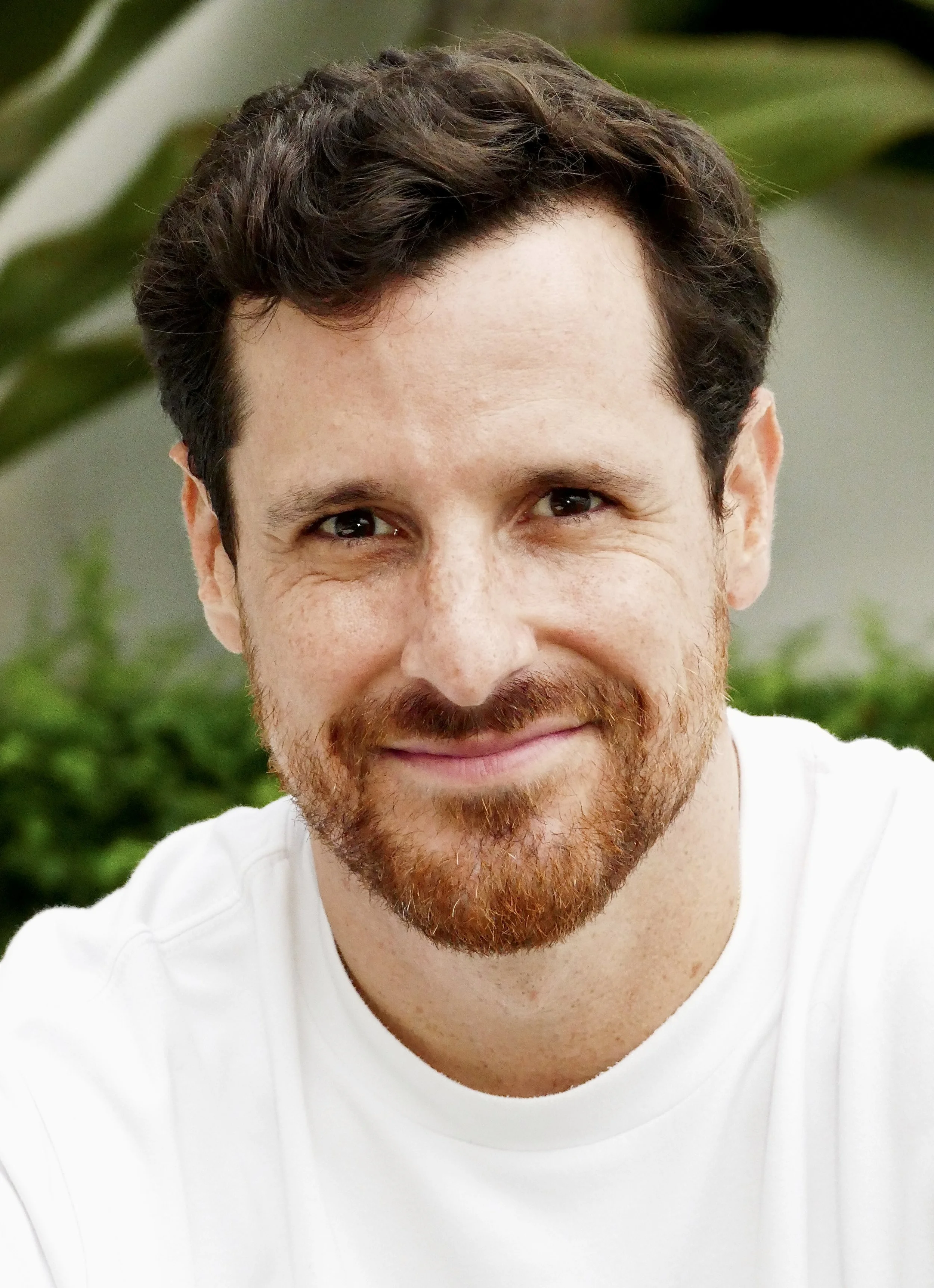 Primer plano de un hombre con barba y cabello castaño, sonriendo, con fondo difuso de plantas verdes.