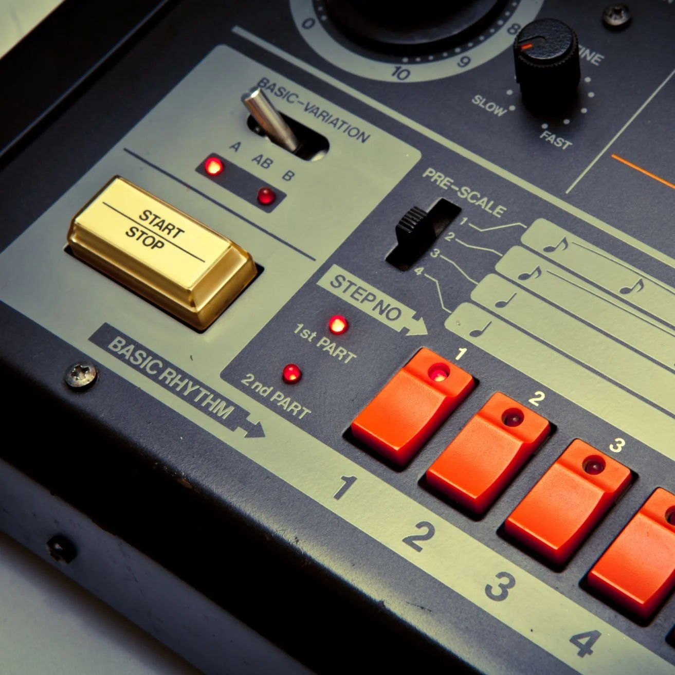Close-up of a vintage electronic music rhythm machine with orange drum pads labeled 1 to 4, a start/stop button, and various control switches and lights.