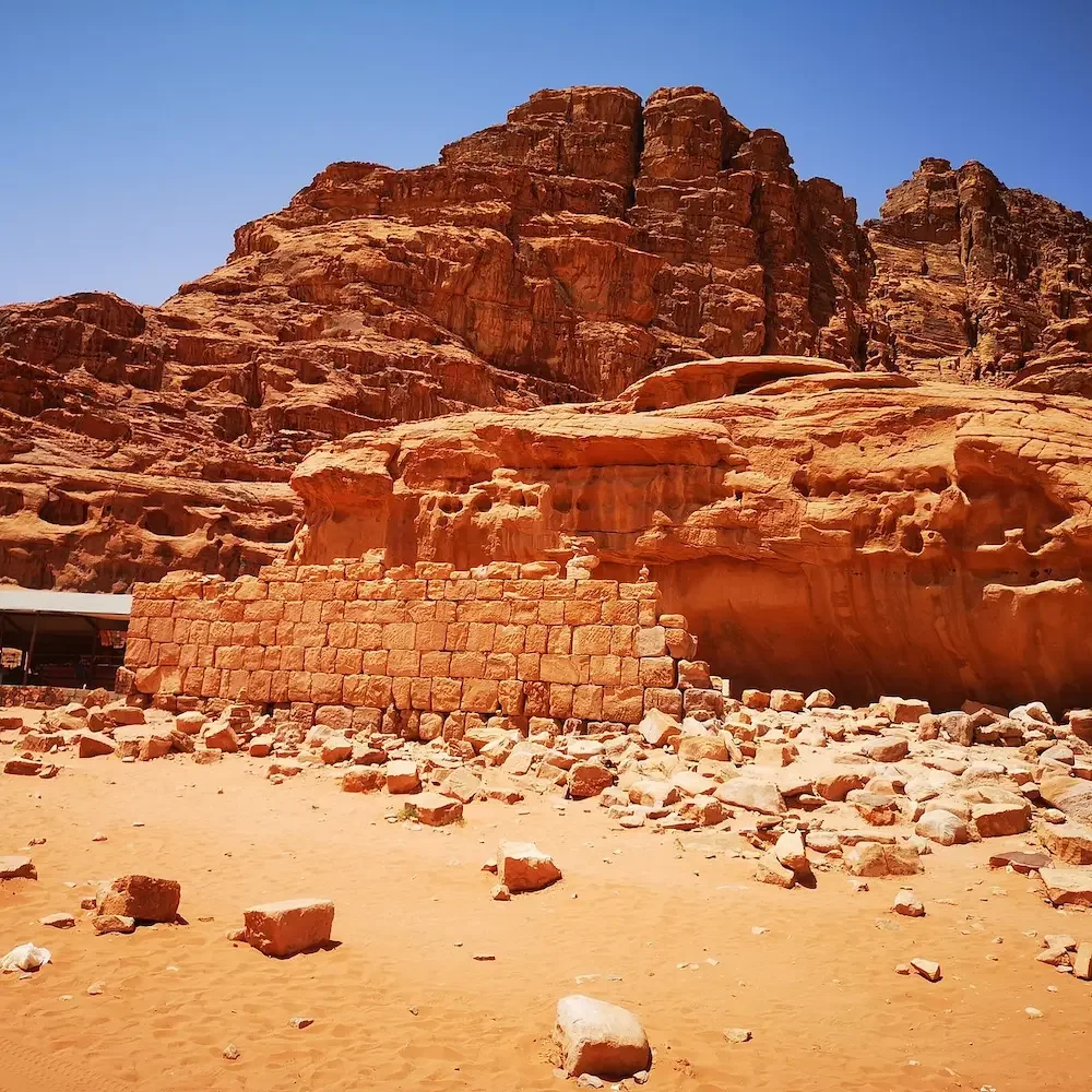 Ancient stone wall built at the base of a red rock formation in a desert landscape under a clear blue sky.