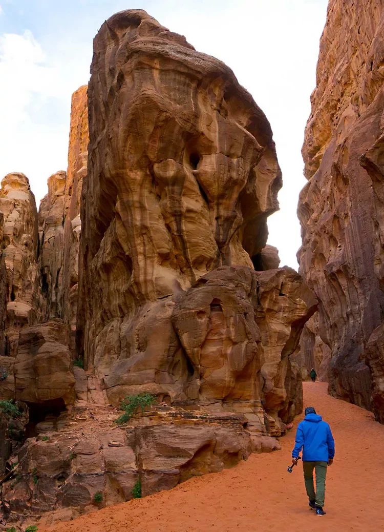 Person in a blue jacket walking along a sandy trail between tall, reddish sandstone rock formations nearby.