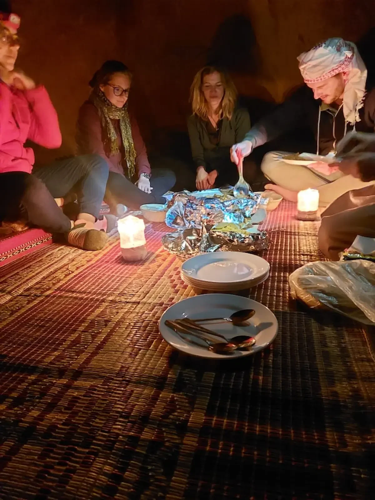 Group of people sitting on a carpeted floor around food items and candles inside a tent.