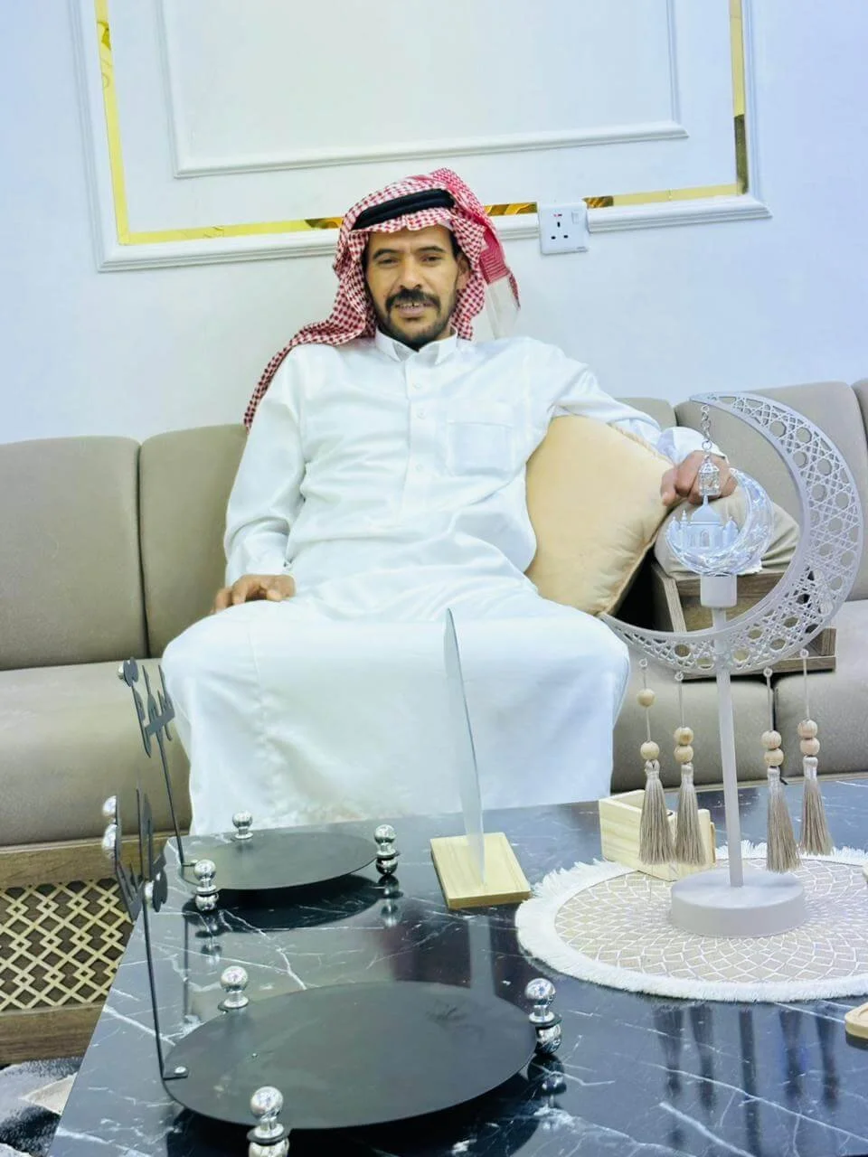 A man dressed in traditional Middle Eastern attire, sitting on a beige sofa, holding a decorative lamp with Islamic design, in a modern living room with a black marble table and decorative objects.