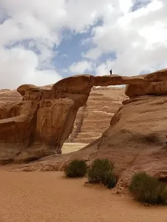 A natural rock arch formation in a desert landscape with a person standing on top, under a partly cloudy sky.