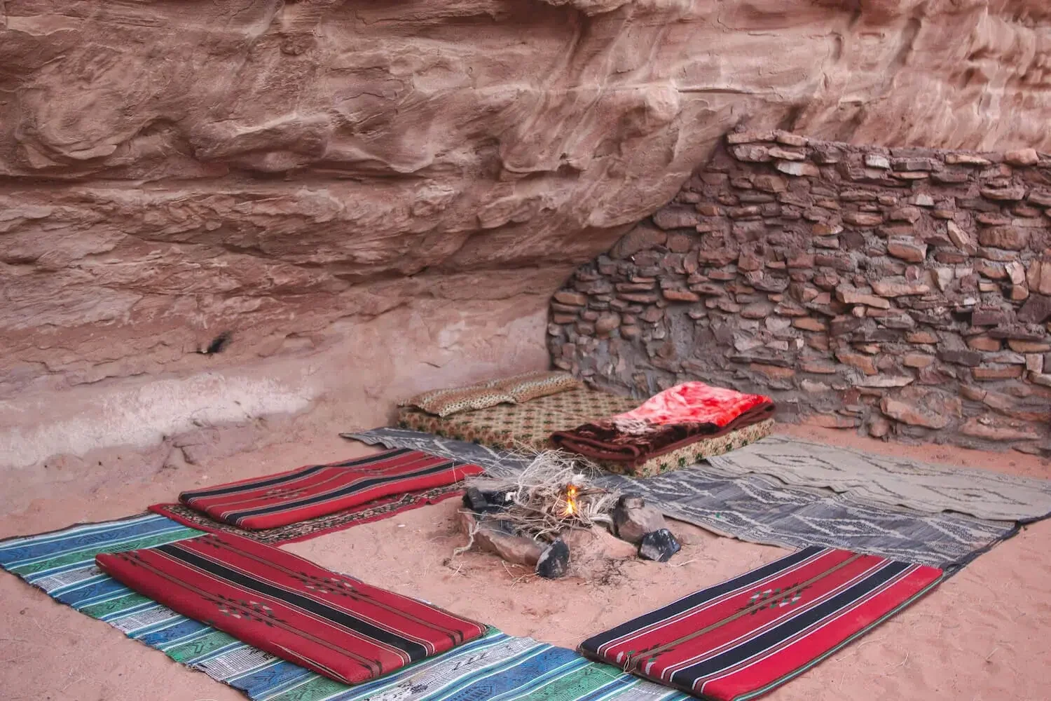 A small outdoor tent shelter with sitting cushions, a fire pit, and blankets, set against a rocky cliff wall, resembling a traditional desert camping setup.