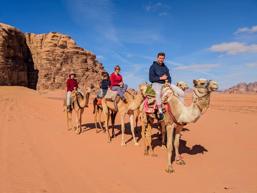 Four people riding camels in a desert with rock formations in the background under a blue sky.