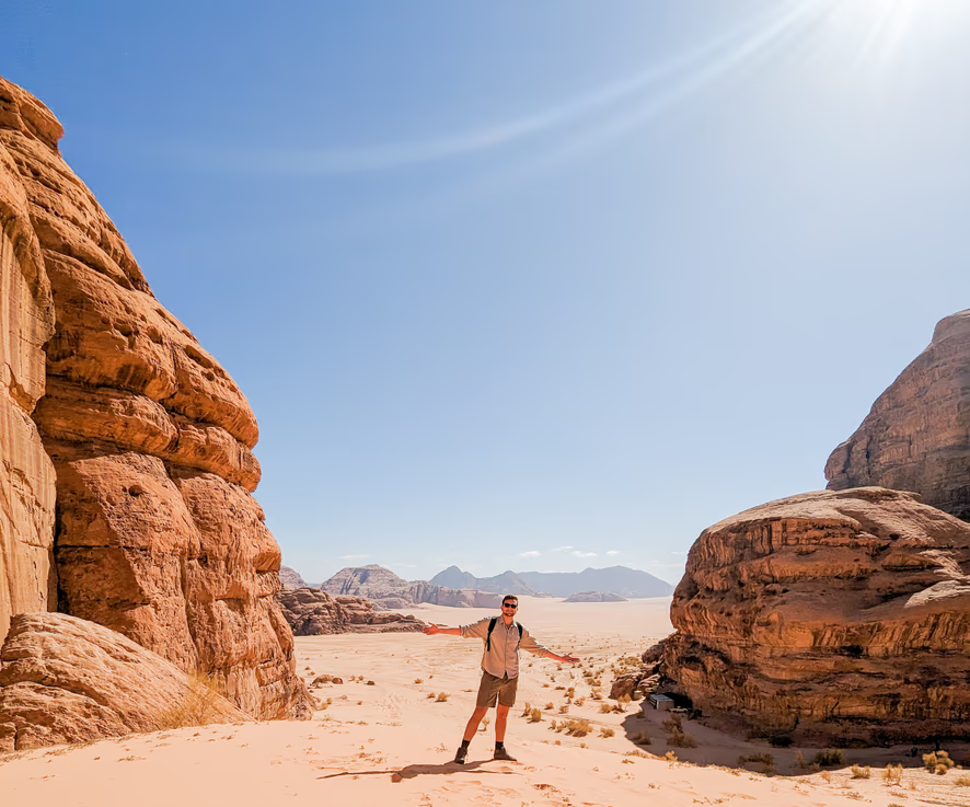 A person standing with arms outstretched in a desert landscape with large rock formations on either side and a clear blue sky overhead.