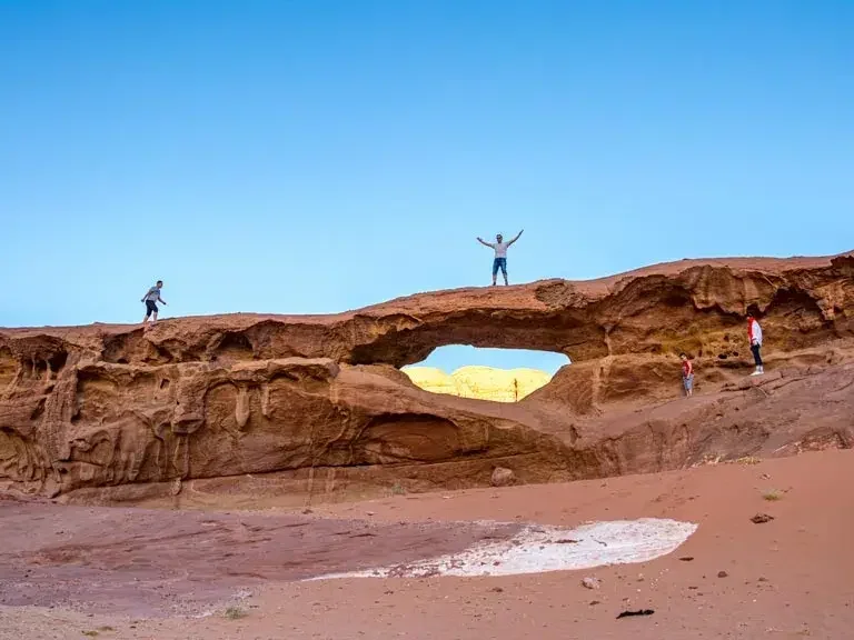 Four people standing on a reddish rock formation with a natural arch and a blue sky background.