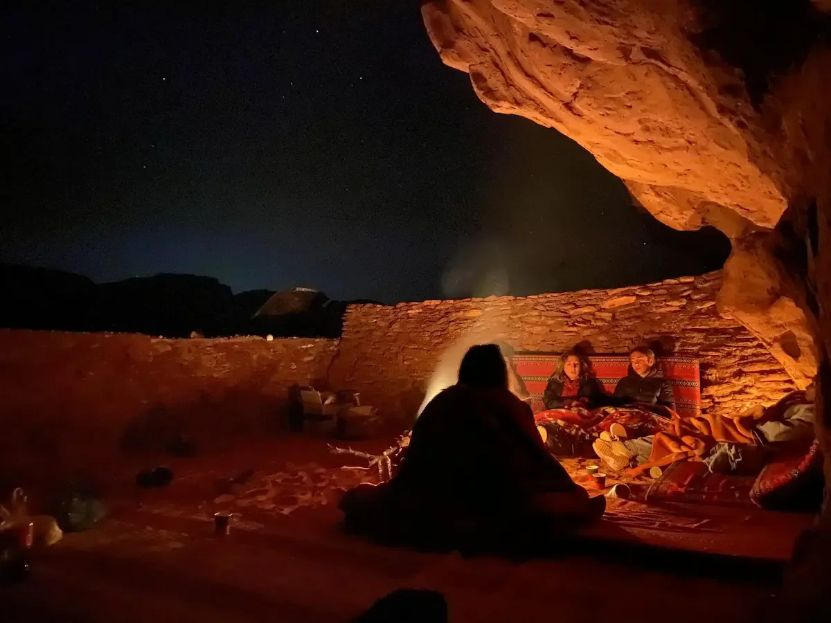 People sitting around a campfire under a starry night sky, inside a cave with reddish rock formations.
