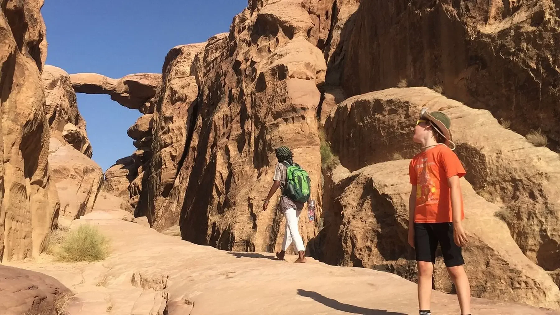 Two children hiking in a desert canyon with large rock formations, one with a green backpack and patterned head covering, the other with a wide-brimmed hat and sunglasses.