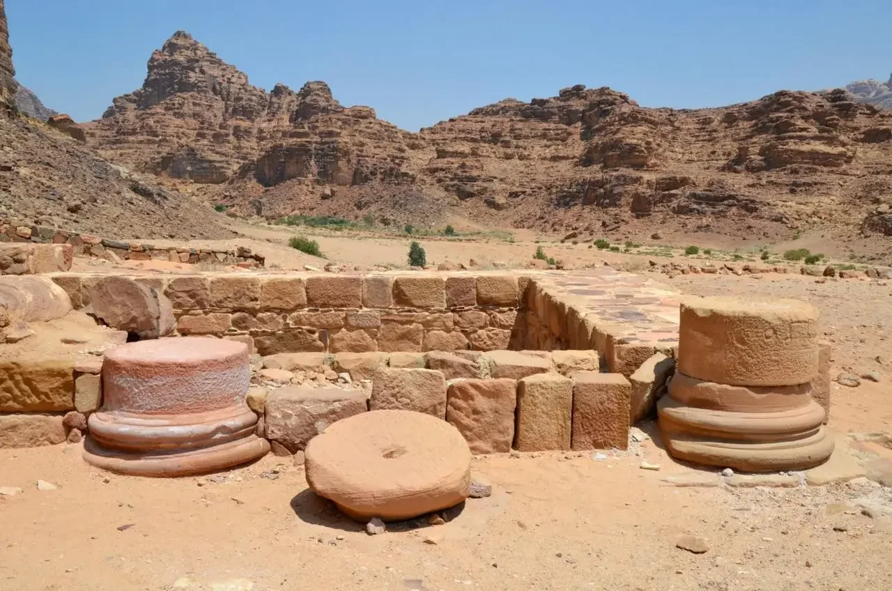 Ancient ruins with stone columns and a partially reconstructed wall set in a desert landscape with mountains in the background.