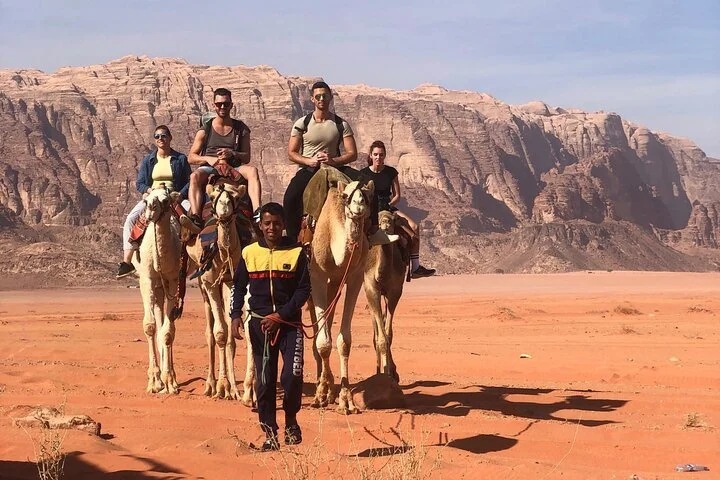 Group of five people riding camels in a desert with red sand, mountains in the background, and a person standing in front holding camel reins.