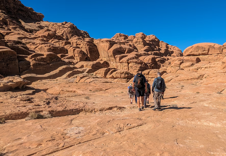 Group of hikers walking through a desert landscape with large rock formations under a clear blue sky.