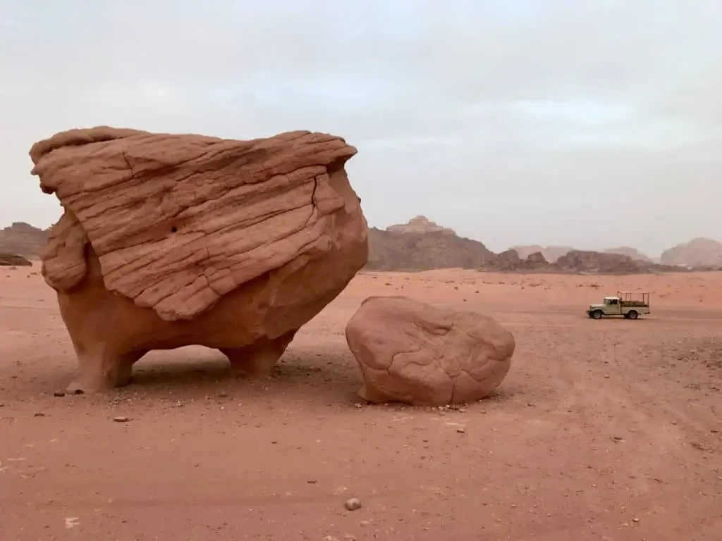 Large reddish rock formations in a desert landscape with a small vehicle in the distance.