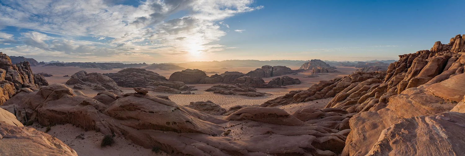 Desert landscape with large reddish rocks and sand dunes under a partly cloudy sky at sunset or sunrise.