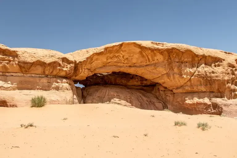 Desert landscape featuring a large natural rock arch formation with sparse vegetation and clear blue sky.