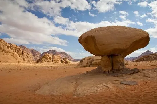 A large mushroom-shaped rock formation in a desert with distant mountains and a partly cloudy sky.