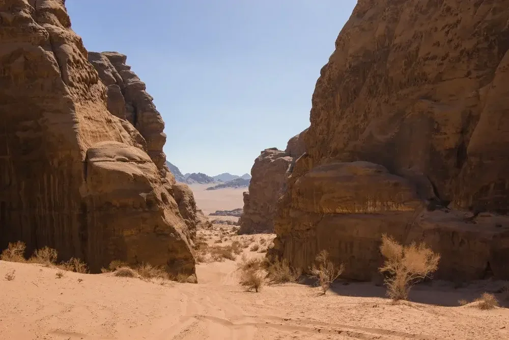Desert landscape with large rock formations on either side and sparse bushes on the sandy ground under a clear blue sky.