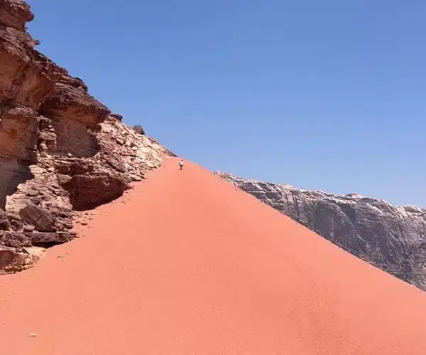 A person hiking on a steep, sandy trail along a rocky mountain slope under a clear blue sky.