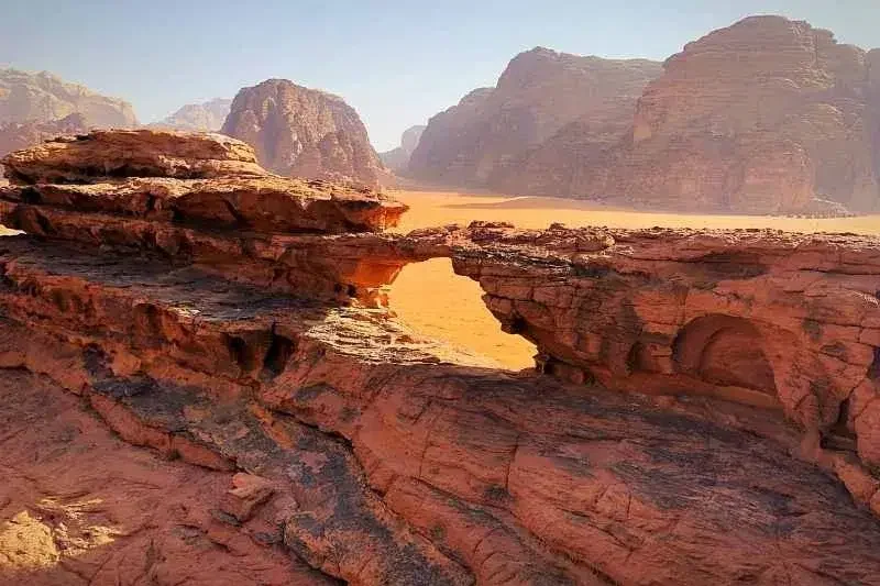 Red rock formations with natural arches in a desert landscape, with mountains in the background under a clear sky.