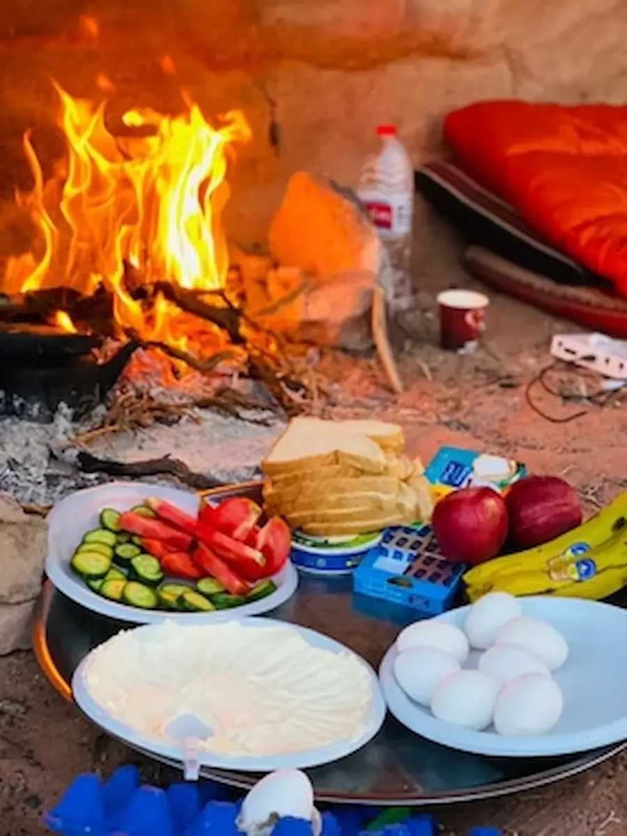 Indoor scene with a campfire, a mattress with a blanket, and a table with various foods including boiled eggs, sliced bread, vegetables, apples, and bananas.