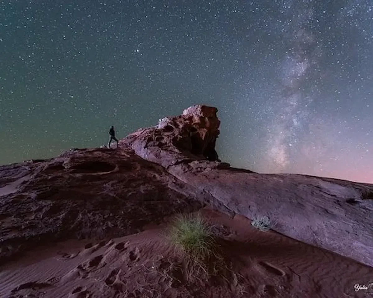 A person standing on a rocky mountain under a starry night sky with the Milky Way galaxy visible.