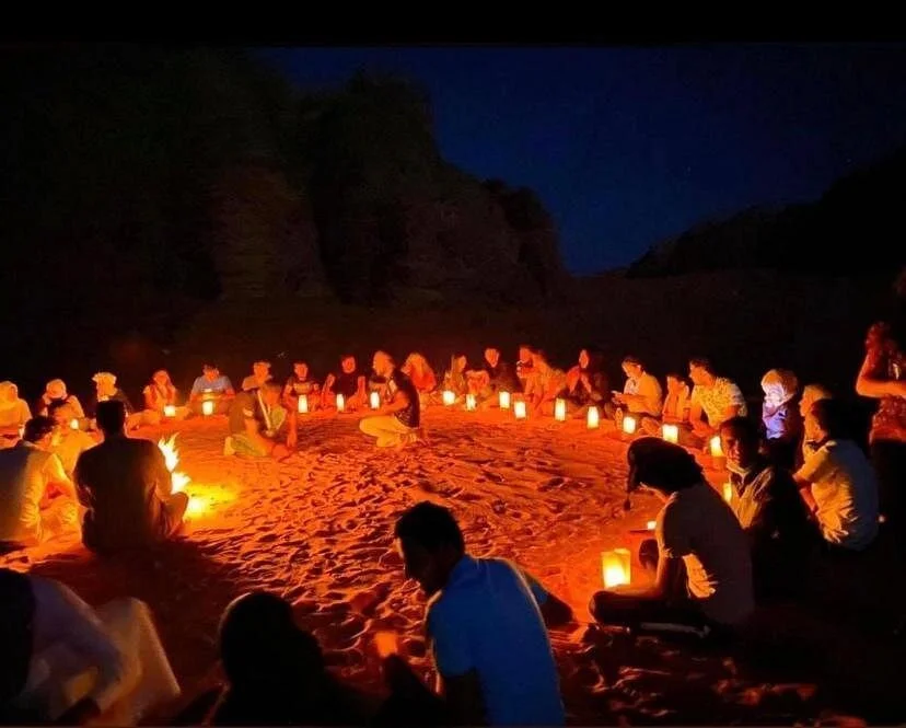 Group of people sitting in a circle on sandy ground at night, illuminated by numerous glowing lanterns