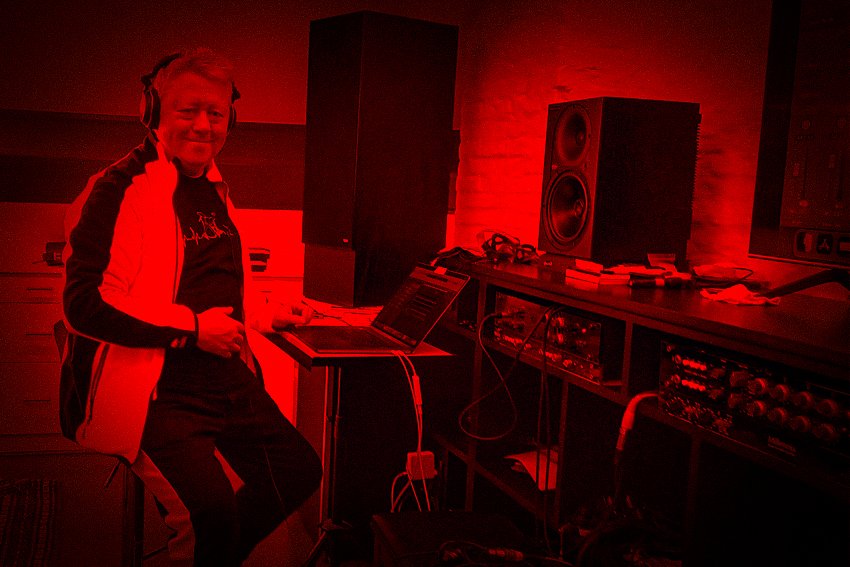 Man in een geluidsstudio met professionele audioapparatuur en luidsprekers, met rode belichting.