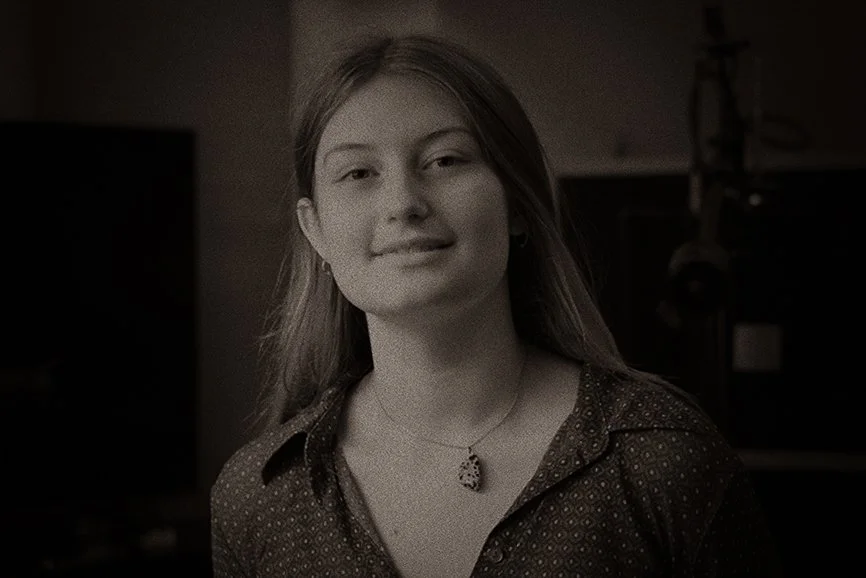 Portret van een jonge vrouw met halflang haar, gekleed in een blouse met kleine patronen, draag een ketting en oorbellen, in een binnenruimte.