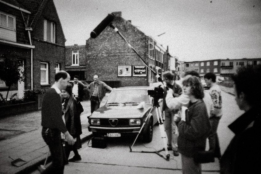Een groep mensen, inclusief een cameraman, staan op een straat bij een oude auto, in een wijk met huizen en een gebouw met een schoorsteen.