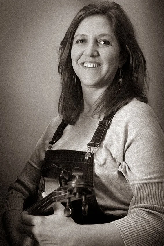 Zwarte en witte portretfoto van een vrouw met schouderlange haar die een gitaar vasthoudt en glimlacht, tegen een effen achtergrond.