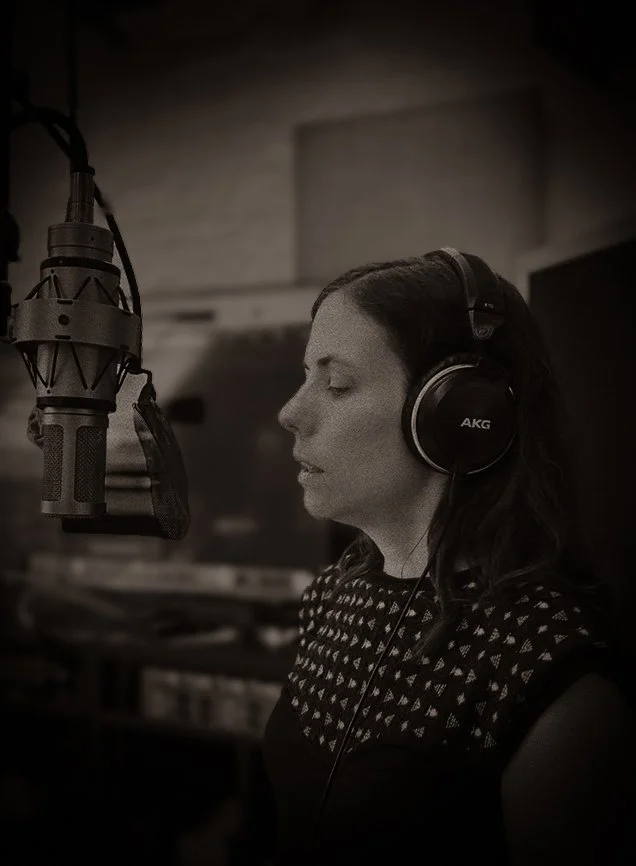 Een vrouw met koptelefoon die in een opname.studio zingt of spreekt voor een microfoon.